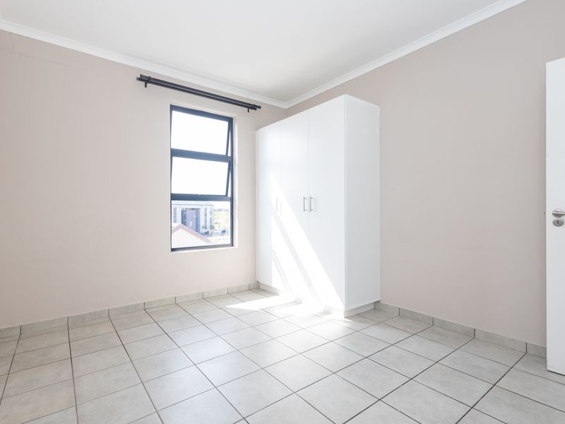 A bright, empty bedroom featuring neutral tiled flooring, a built-in white wardrobe, and a window with an aluminium frame.