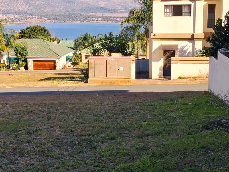 A view of a vacant residential plot featuring a boundary wall and an electrical utility box, with surrounding neighborhood and water views.