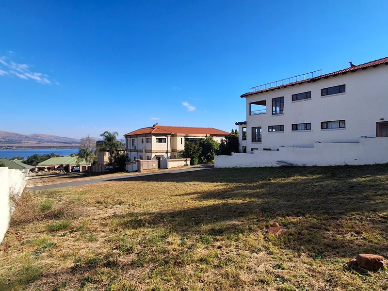 A view of a vacant residential plot featuring a boundary wall, adjacent multi-story homes, and distant water views.