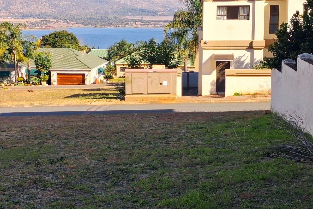 A view of a vacant residential plot featuring a boundary wall and an electrical utility box, with surrounding neighborhood and water views.
