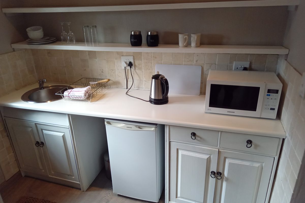 A compact kitchenette featuring white cabinetry, a stainless steel sink, tiled backsplash, and open shelving for storage.