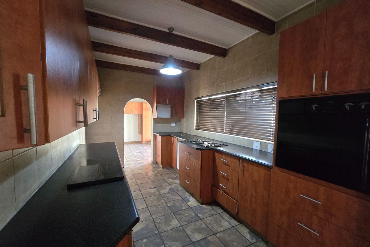 A functional kitchen featuring wood-tone cabinetry, dark countertops, built-in oven, gas hob, and exposed timber ceiling beams with tiled flooring throughout.