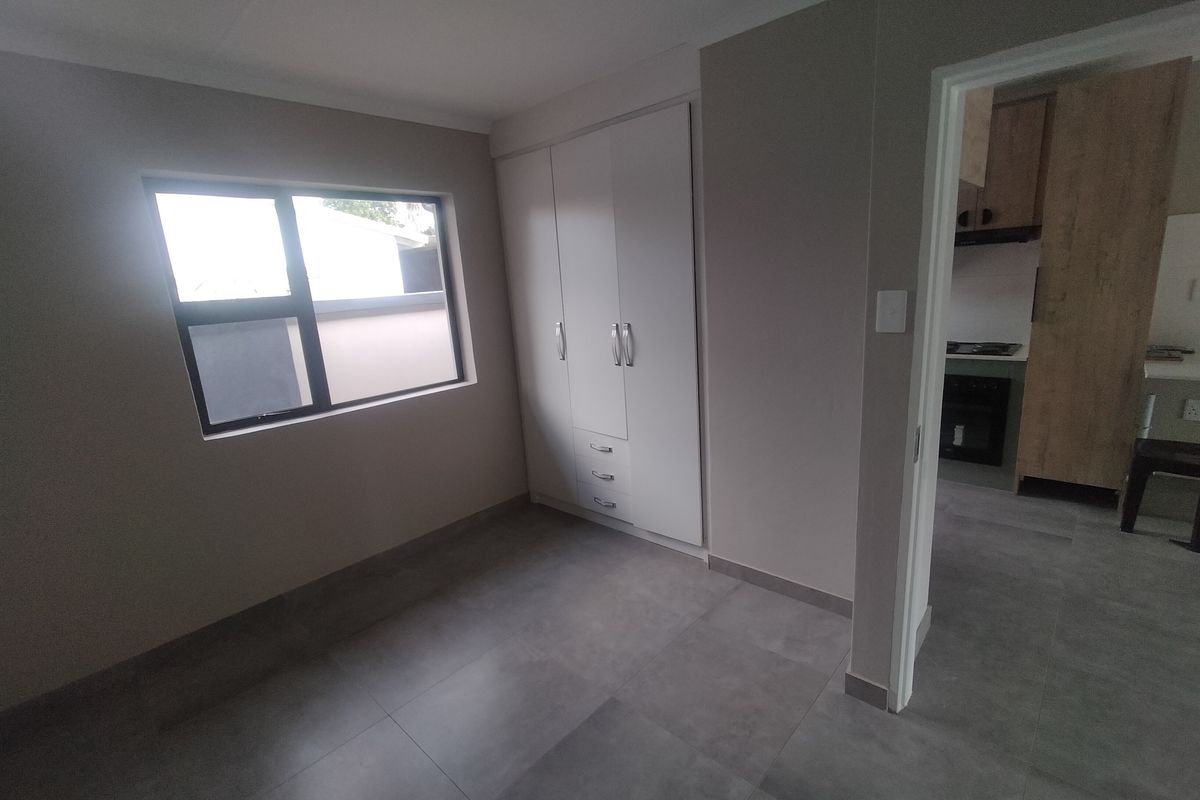 A clean, modern bedroom featuring neutral walls, large grey floor tiles, and built-in cupboards, with a view into the adjacent kitchen area.
