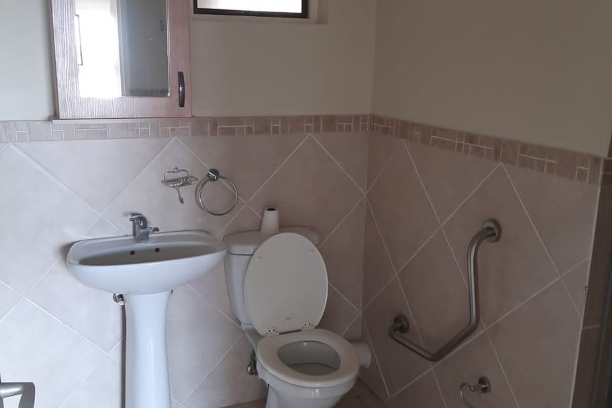 A functional bathroom featuring a pedestal basin, toilet, wall-mounted grab rails, and neutral ceramic wall and floor tiling.