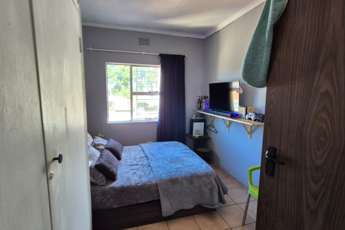 A compact bedroom featuring tiled flooring, a built-in cupboard, and a window with security bars, providing a functional living space.