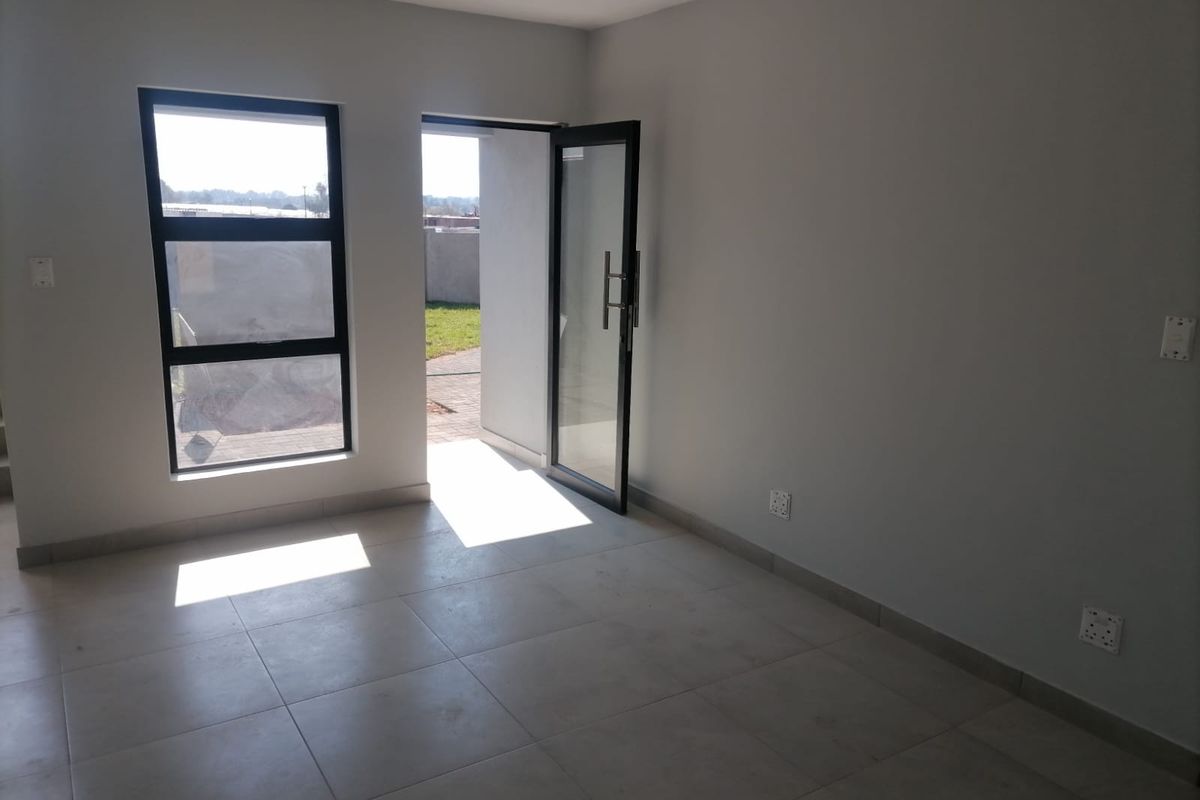 A bright, empty living area featuring neutral floor tiles, modern aluminium window frames, and a glass door providing direct access to the outdoor space.