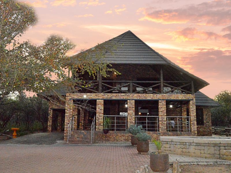 A charming double-story brick house with a thatched roof, featuring a spacious covered patio and a paved driveway, set in a natural bushveld environment.