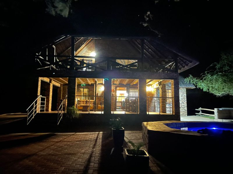 Night view of a double-story brick house featuring a thatched roof, covered patio, balcony, and a swimming pool in the foreground.