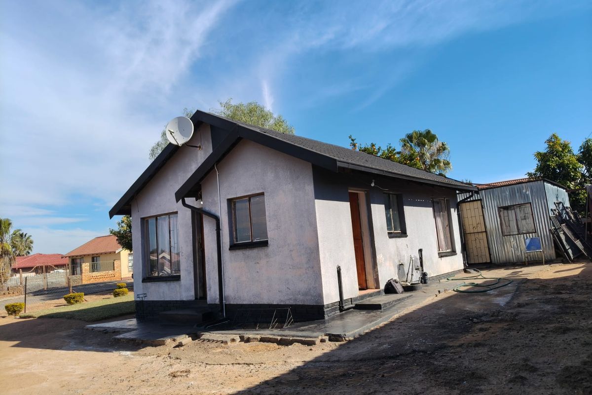 Rear view of a detached house featuring a metal roof, satellite dish, and concrete apron, set on a dirt-surfaced plot.