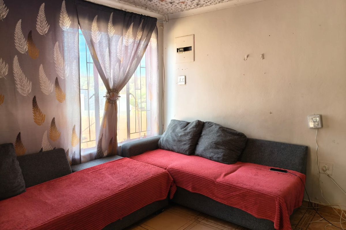 A living room area featuring tiled flooring, security bars on the window, and an exposed electrical distribution board, requiring cosmetic updates to the ceiling and walls.