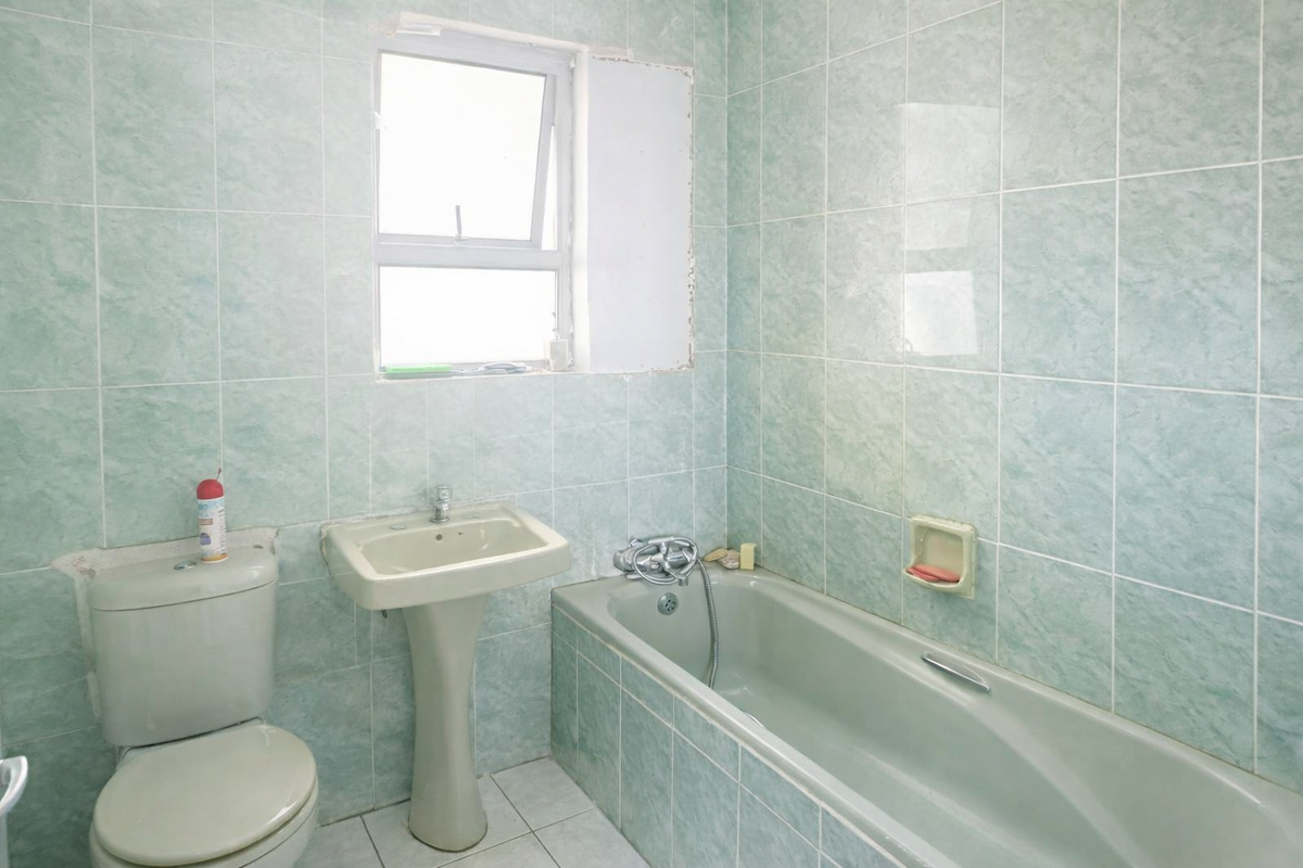 A dated bathroom featuring a pedestal basin, bathtub, and toilet with light green wall tiling and an unfinished window surround.