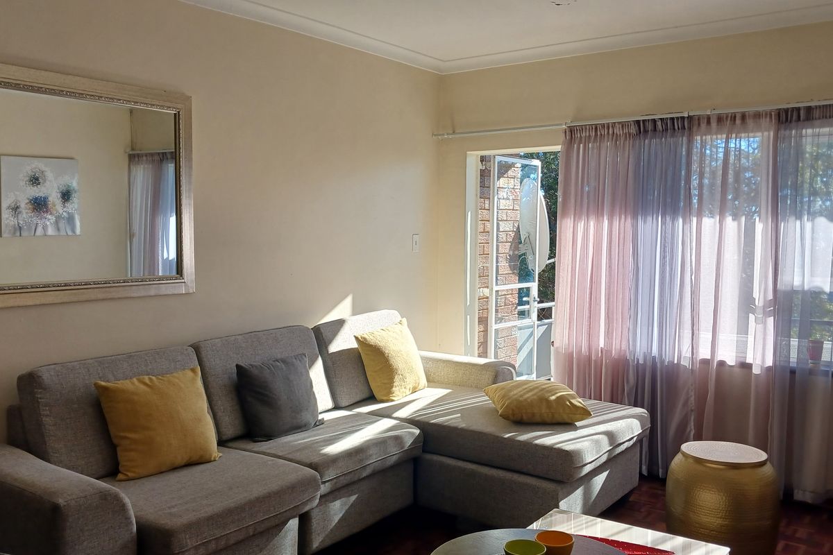 A bright living room featuring parquet flooring, large windows with sheer curtains, and direct access to an outdoor balcony area.