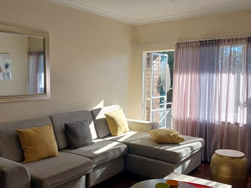 A bright living room featuring parquet flooring, large windows with sheer curtains, and direct access to an outdoor balcony area.