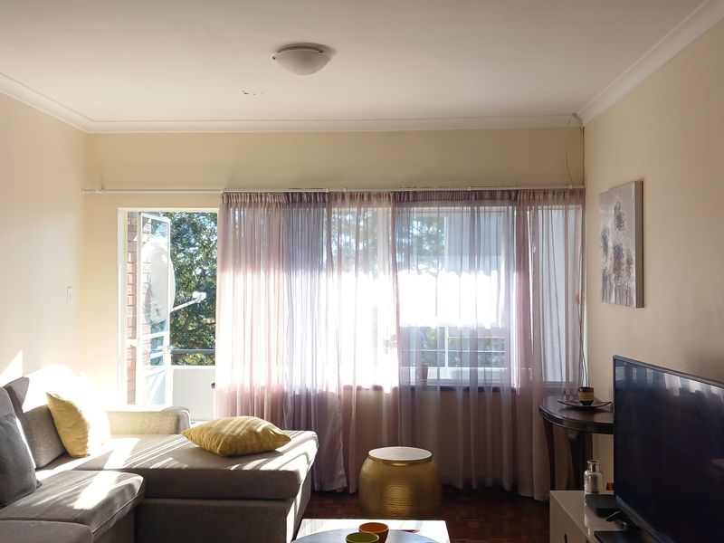 A bright living room featuring parquet flooring, large windows with sheer curtains, and direct access to an outdoor balcony area.