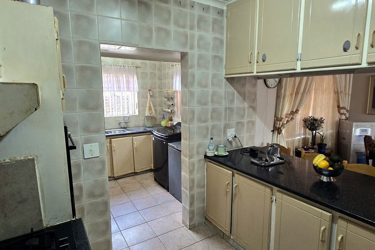 A dated kitchen featuring built-in cabinetry, granite countertops, and ceramic tiling, with an adjoining scullery area visible through the doorway.