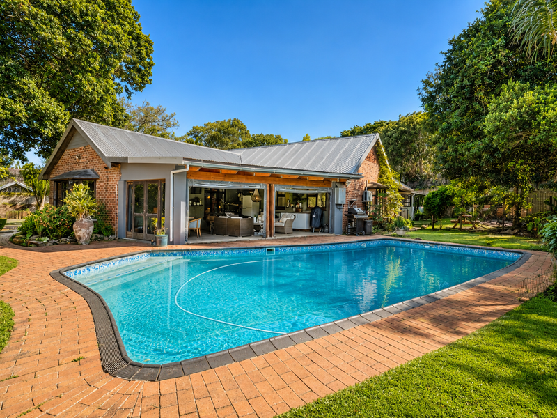 A well-maintained swimming pool area featuring brick paving, a landscaped garden, and a covered patio attached to the main residence.