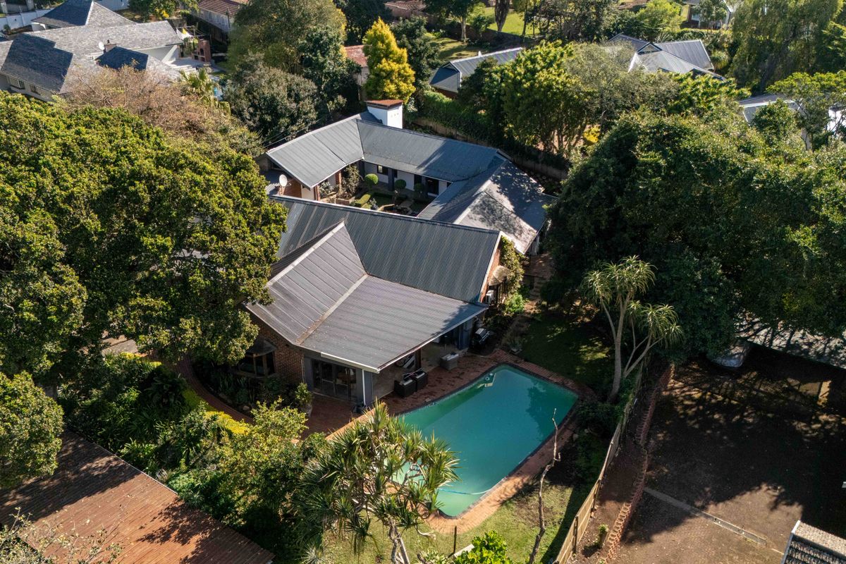 Aerial view of a residential property featuring a swimming pool, covered patio, and mature garden surroundings with a metal roof structure.