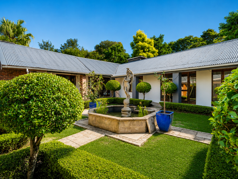 A well-maintained rear courtyard featuring a central stone fountain, manicured hedging, paved pathways, and a classic corrugated metal roof structure.