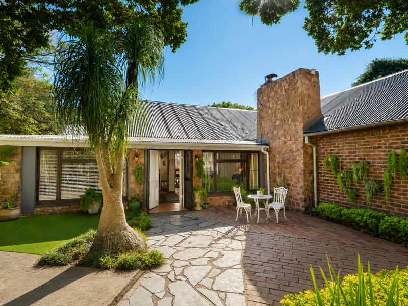 A charming rear exterior view featuring a stone chimney, corrugated metal roof, and a combination of stone and brick paved patio areas.