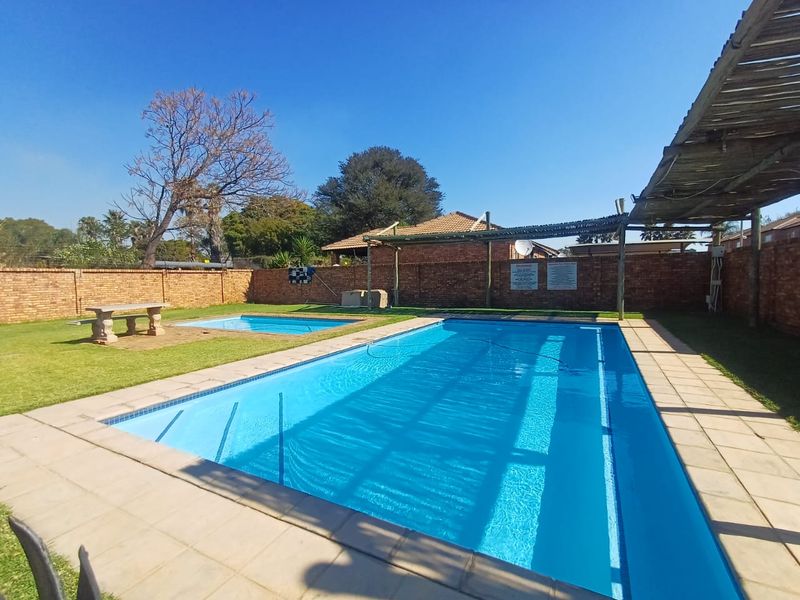 Communal swimming pool area featuring a large main pool, a smaller wading pool, paved surrounds, and a timber pergola structure enclosed by brick walls.