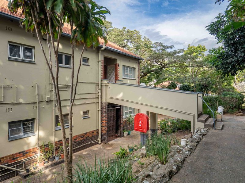 Rear view of the flat complex featuring a raised concrete walkway, brick and plaster facade, and established garden surroundings.