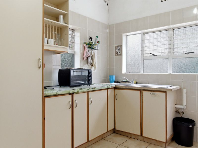 A functional kitchen featuring dated cabinetry, laminate countertops, a stainless steel sink, and tiled splashbacks with louvered windows for natural ventilation.