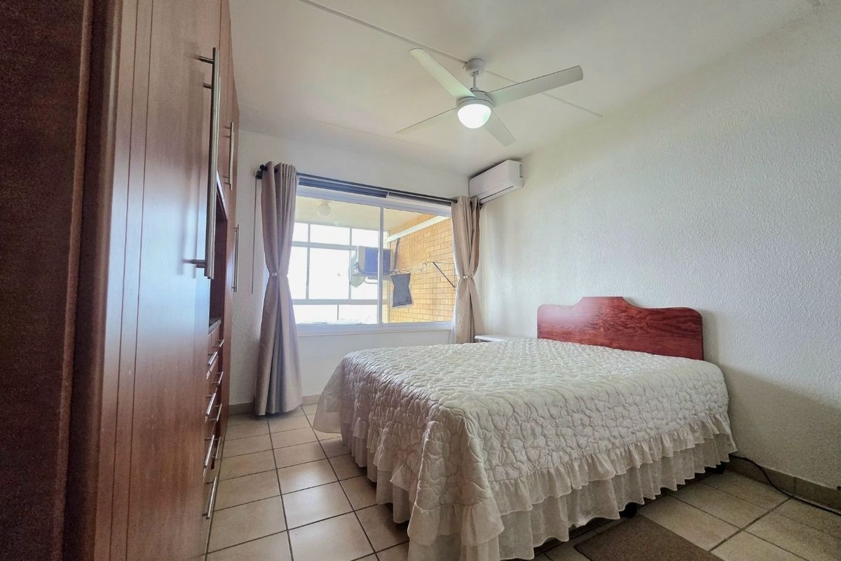 A bedroom featuring built-in cupboards, a ceiling fan, and an air conditioning unit, with tiled flooring and natural light from the window.