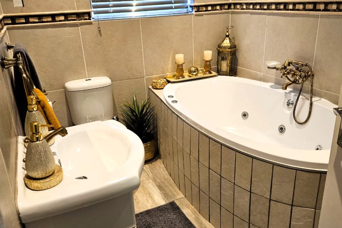 A well-maintained bathroom featuring a corner jetted bathtub, pedestal washbasin, toilet, and neutral wall and floor tiling with decorative accents.