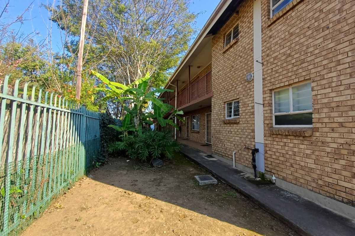 Rear exterior view of the apartment building featuring a brick facade, concrete walkway, and metal security fencing along the perimeter.
