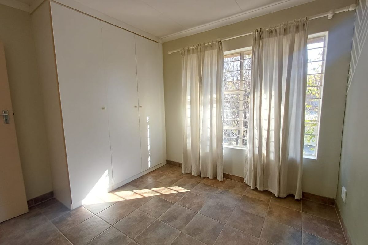 A bright, empty bedroom featuring neutral tiled flooring, built-in cupboards, and windows fitted with security bars.