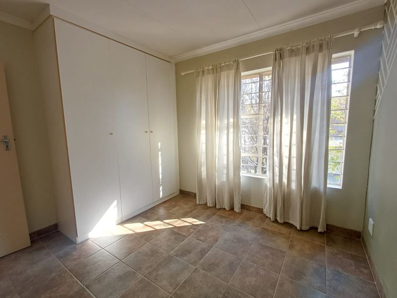 A bright, empty bedroom featuring neutral tiled flooring, built-in cupboards, and windows fitted with security bars.