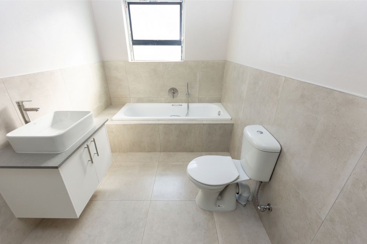 A clean, modern bathroom featuring a built-in bathtub, wall-mounted basin with vanity, and a toilet, finished with neutral floor-to-ceiling tiling.