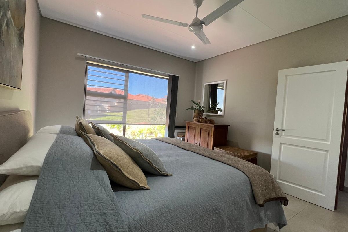 A well-maintained bedroom featuring neutral wall tones, tiled flooring, a modern ceiling fan, and large windows with roller blinds.