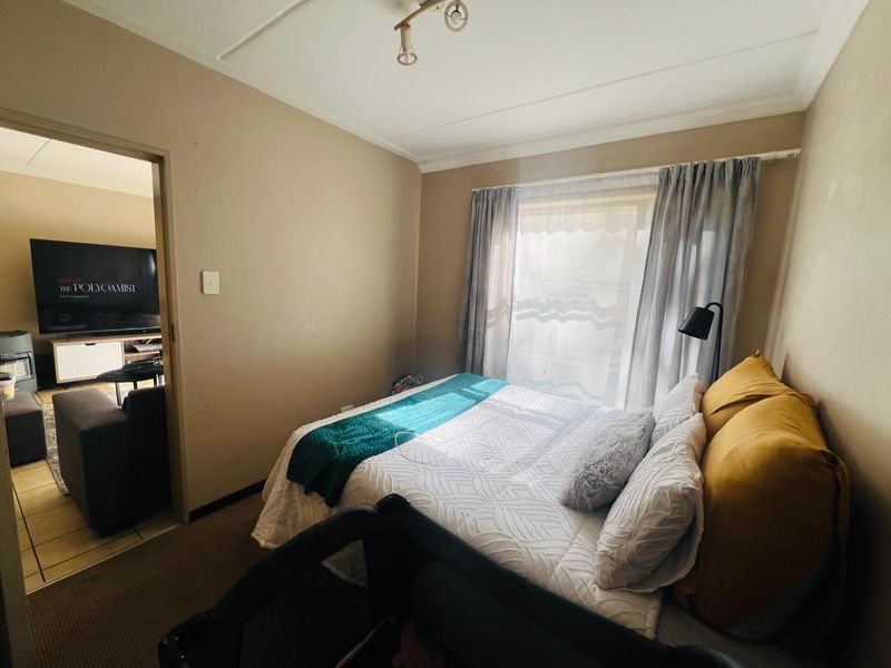 A functional bedroom featuring neutral wall tones, a window with curtain rails, and overhead lighting, with an adjacent view into the living area.