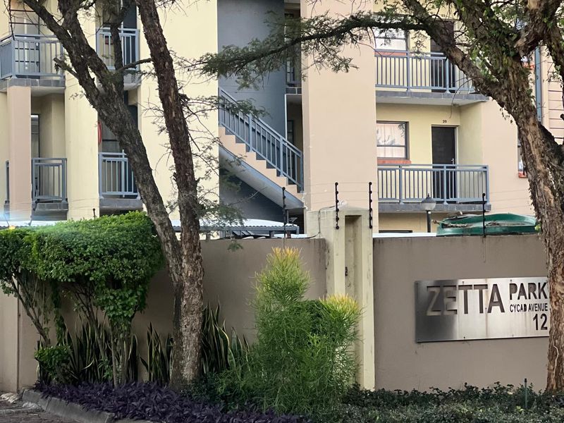 Front exterior view of Zetta Park apartment complex featuring a paved entrance, security fencing, and multi-level residential units with balconies.