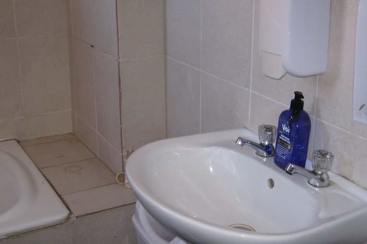A functional bathroom featuring a pedestal basin and a built-in bathtub with tiled surrounds, showing signs of wear and dated finishes.