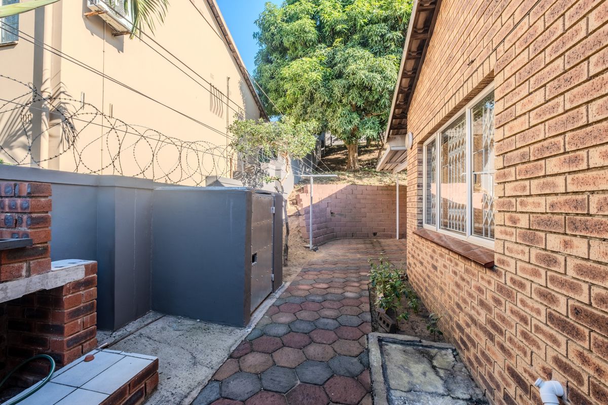 Rear exterior area featuring a brick braai, paved walkway, and retaining wall, secured with perimeter razor wire and window security bars.