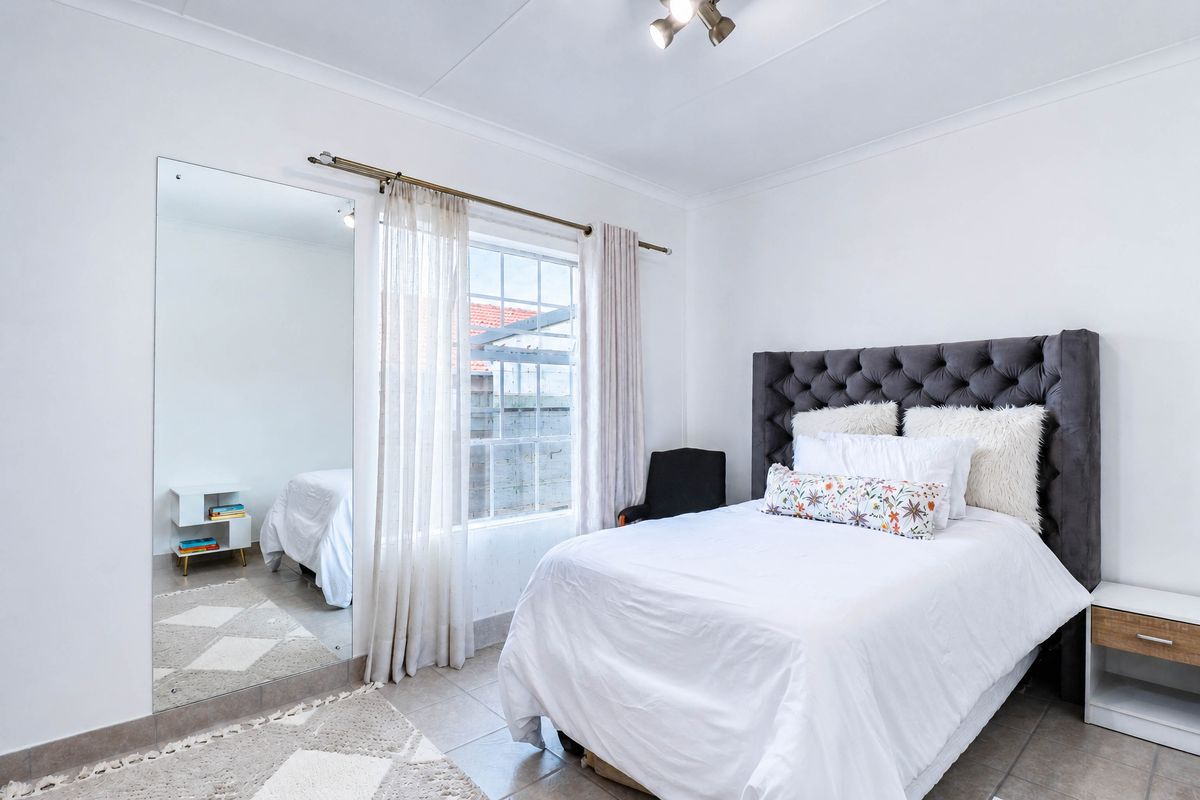A well-lit bedroom featuring tiled flooring, a large wall-mounted mirror, and a window with curtain fixtures, providing a clean and functional living space.