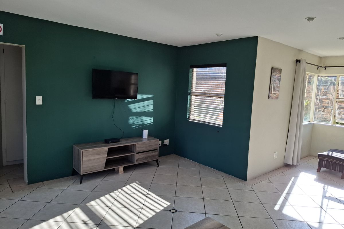 A bright living room featuring ceramic floor tiles, recessed downlighting, and large windows with venetian blinds, finished with a bold green accent wall.