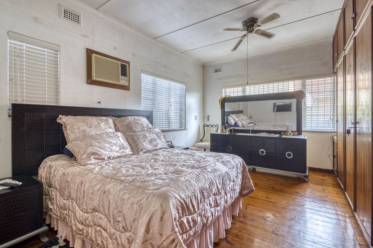 Spacious bedroom featuring timber flooring, built-in wardrobes, a ceiling fan, and a window-mounted air conditioning unit.