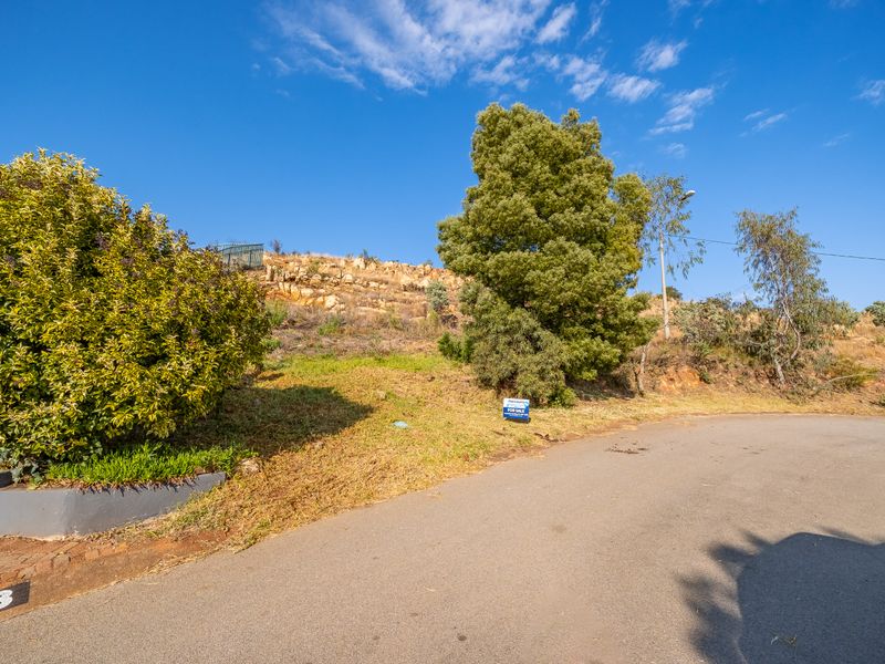A view of a vacant residential plot featuring a sloped landscape, mature trees, and a retaining wall, situated along an asphalt street.