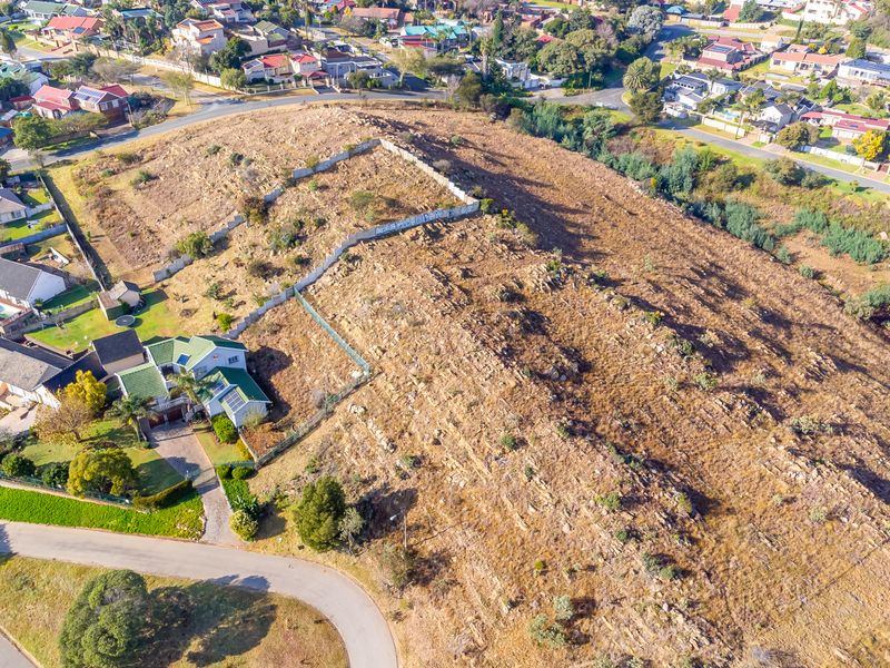 An aerial view of a vacant residential plot featuring natural rocky terrain and boundary fencing, situated within an established suburban neighborhood.