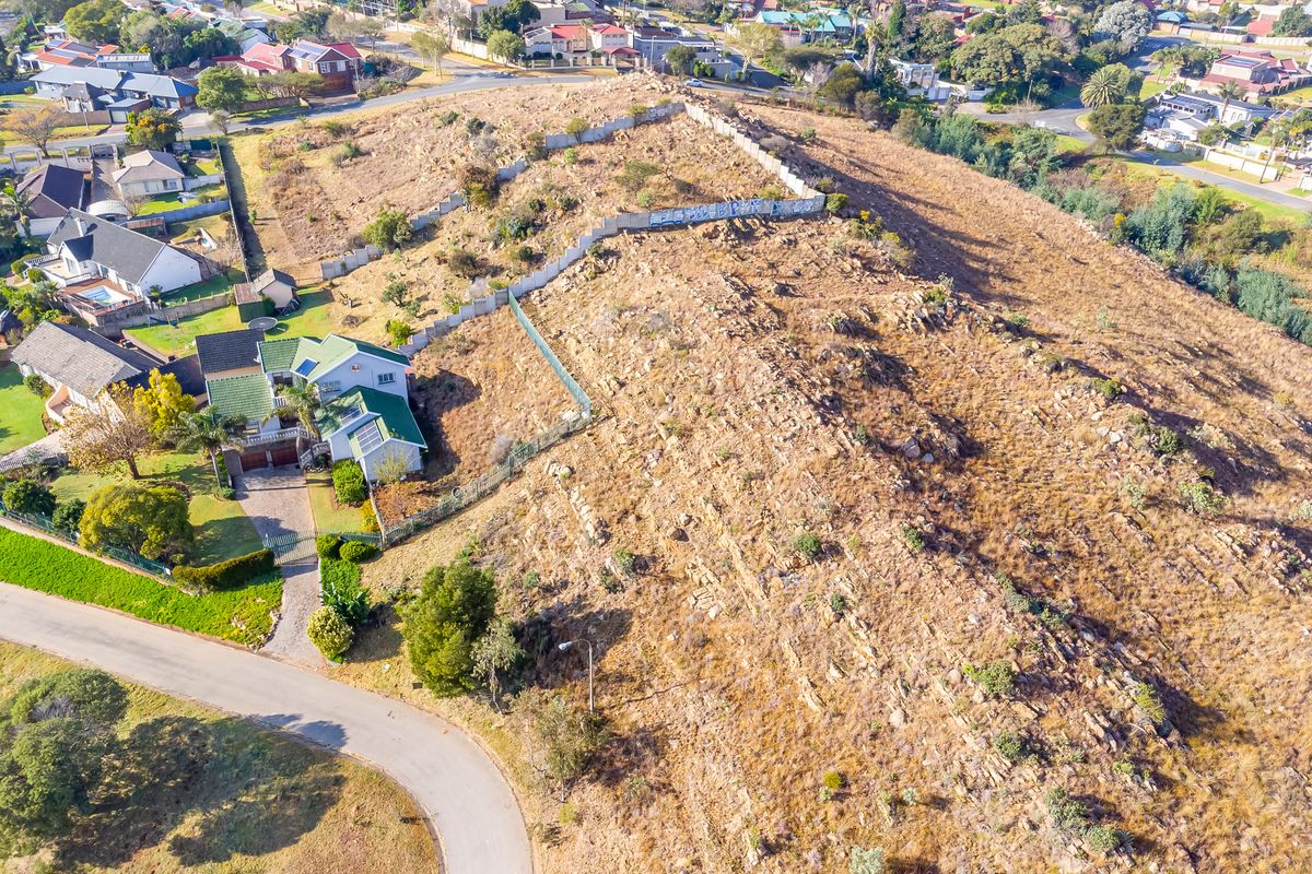 Aerial view of a vacant residential plot featuring natural rocky terrain, perimeter fencing, and a retaining wall in a suburban neighborhood.