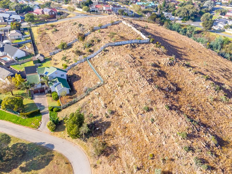 Aerial view of a vacant residential plot featuring natural rocky terrain, perimeter fencing, and a retaining wall in a suburban neighborhood.