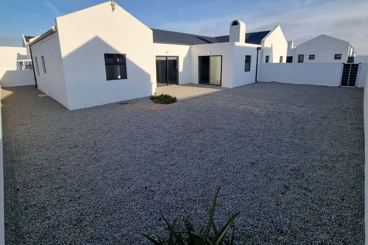 A spacious rear exterior view of a modern white house featuring a gravel yard, boundary walls, and sliding glass door access.