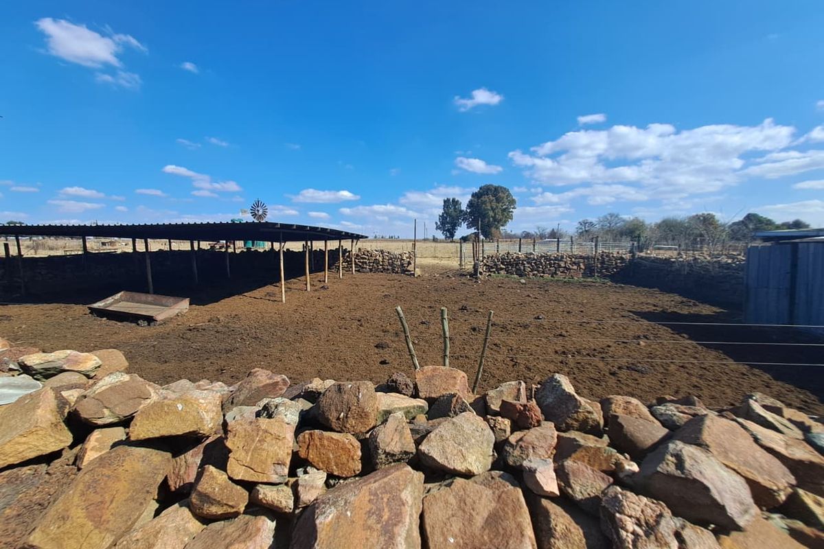 An outdoor farm enclosure featuring stone boundary walls, a large open-sided livestock shelter, and a traditional windmill structure in a rural setting.