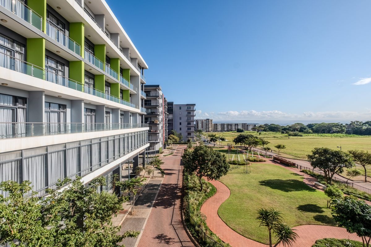 A view of the apartment complex exterior featuring modern glass balconies, paved pathways, and well-maintained landscaped gardens overlooking an open green space.
