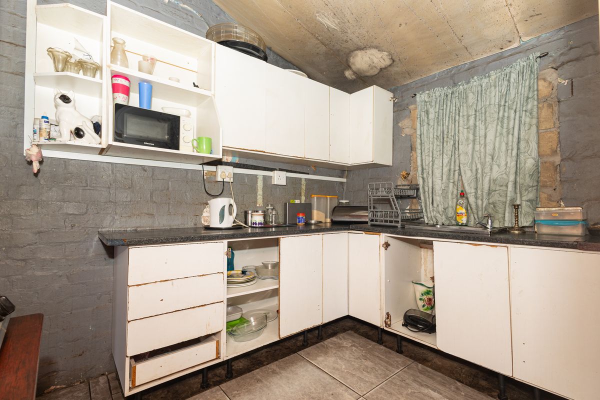 A dated kitchen featuring exposed brick walls, basic cabinetry, and tiled flooring, requiring significant renovation and cosmetic updates to improve functionality and aesthetic appeal.