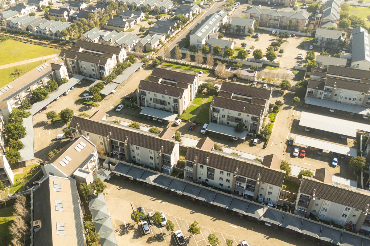 Aerial view of a residential apartment complex featuring multiple buildings, paved driveways, covered parking structures, and well-maintained communal garden areas.