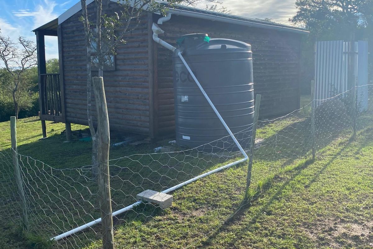 Rear view of a timber cabin featuring a water storage tank, guttering system, and wire mesh perimeter fencing on a grassy plot.